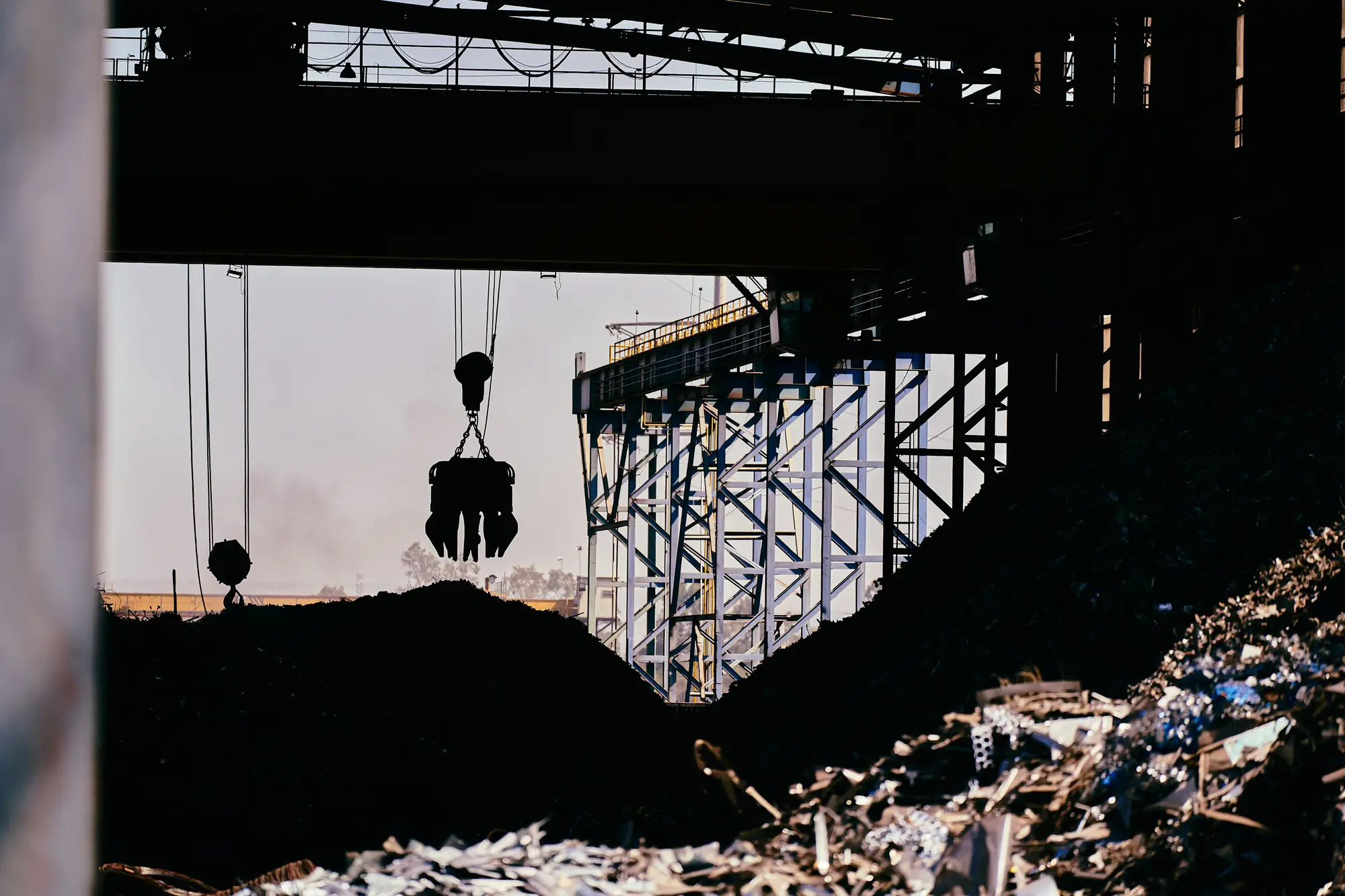 A grab hanging from an overhead gantry above a dark scrap stockpile, seen from the yard floor